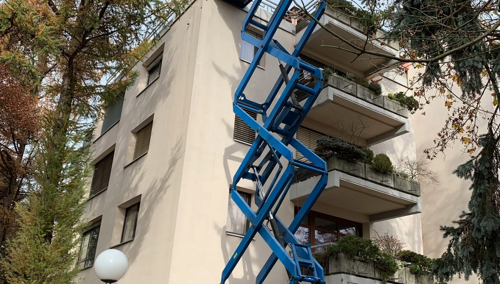 Scherenarbeitsbühne vor einem bewohnten Wohnhaus mit bepflanzten Terrassen.