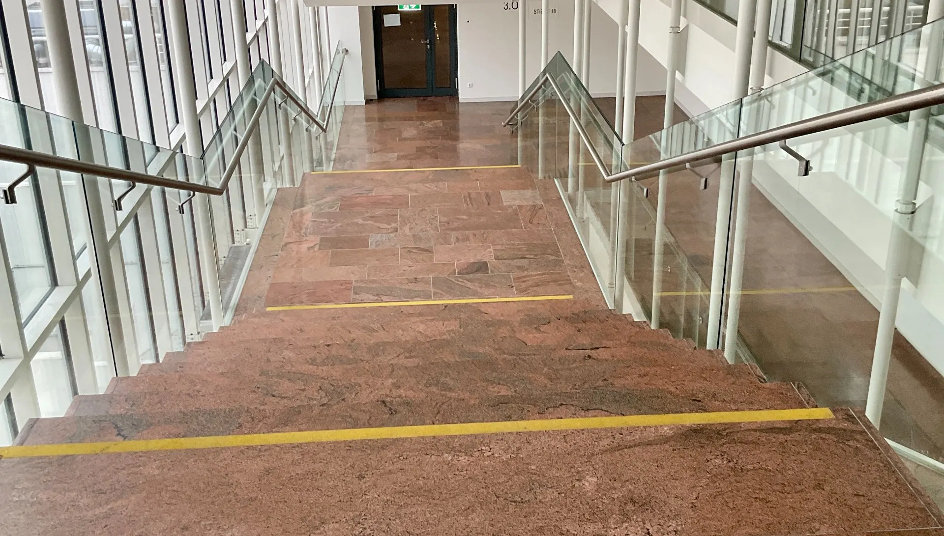 Treppenanlage mit Glasbrüstungen und Steinboden in einem Bürogebäude.