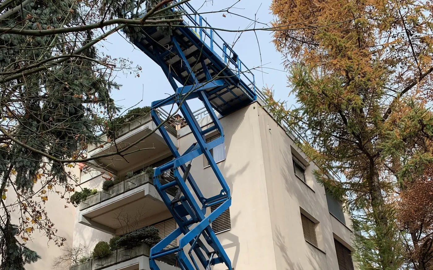 Ausgefahrene Scherenarbeitsbühne mit Arbeitskorb an einem bewohnten Wohnhaus mit bepflanzten Terrassen.