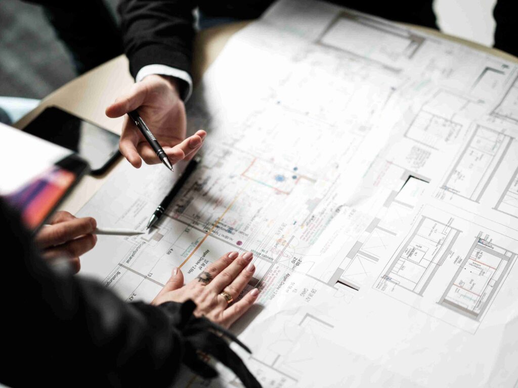 H&auml;nde von Experten der Themis Baumanagement bei einer Besprechung &uuml;ber einem detaillierten architektonischen Grundrissplan auf einem Schreibtisch.