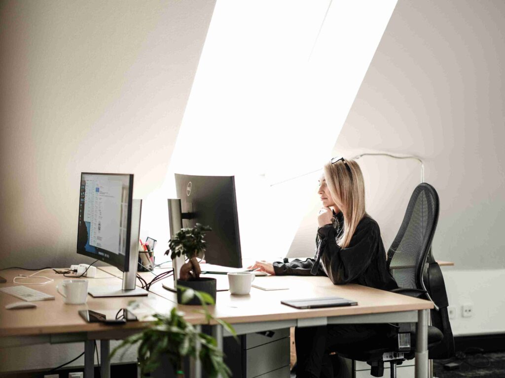 Eine Mitarbeiterin von Themis sitzt konzentriert am Schreibtisch vor zwei Computerbildschirmen in einem hellen Büro und plant ein Bauprojekt.