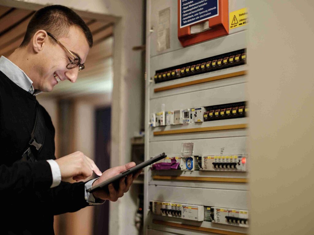 Ein lächelnder Prüfingenieur von Themis dokumentiert mit einem Tablet den Zustand eines Elektro-Verteilerkastens bei einer Objektsicherheitsprüfung im Gebäude.