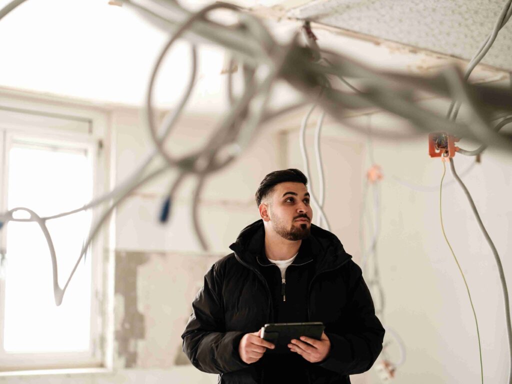 Ein Bauleiter von Themis begutachtet mit einem Tablet eine Baustelle im Innenbereich mit freiliegenden Kabelsträngen an der Decke.